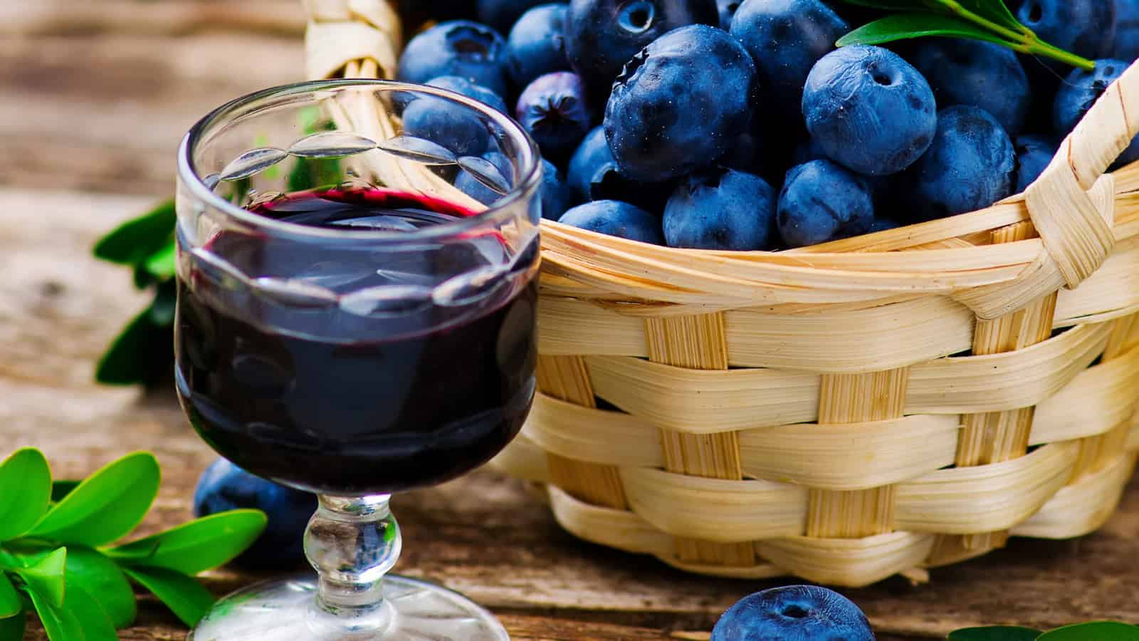 A glass of blueberry juice is placed on a wooden surface next to a wicker basket filled with fresh blueberries. Some green leaves are scattered around the basket.