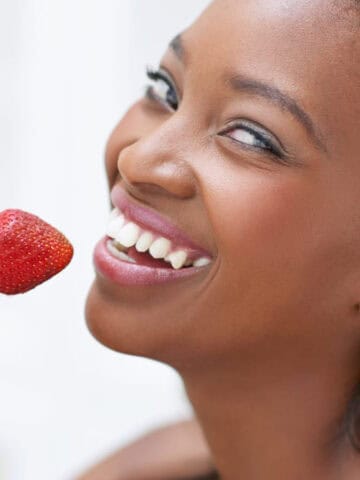 A woman with long dark hair and a joyful expression is holding a strawberry near her face. She is smiling widely and appears to be about to eat the fruit. The background is blurred and white, focusing attention on her face and the strawberry.