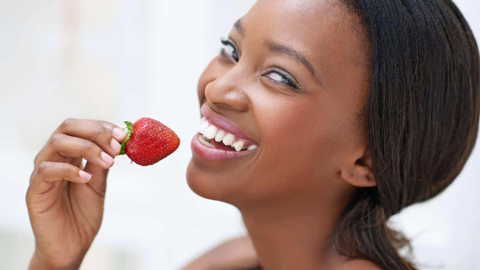 A woman with long dark hair and a joyful expression is holding a strawberry near her face. She is smiling widely and appears to be about to eat the fruit. The background is blurred and white, focusing attention on her face and the strawberry.
