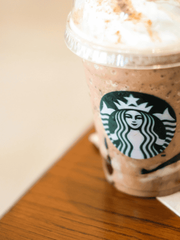 A Starbucks frappuccino topped with whipped cream sits on a wooden table next to a Starbucks-branded napkin. The cup features the Starbucks logo, and the beverage appears to be a blended iced drink with chocolate and cream.