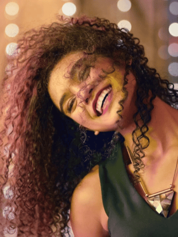 A person with curly hair is smiling with their head tilted back in front of a blurred background with bokeh lights. They are wearing a sleeveless top and a geometric necklace.