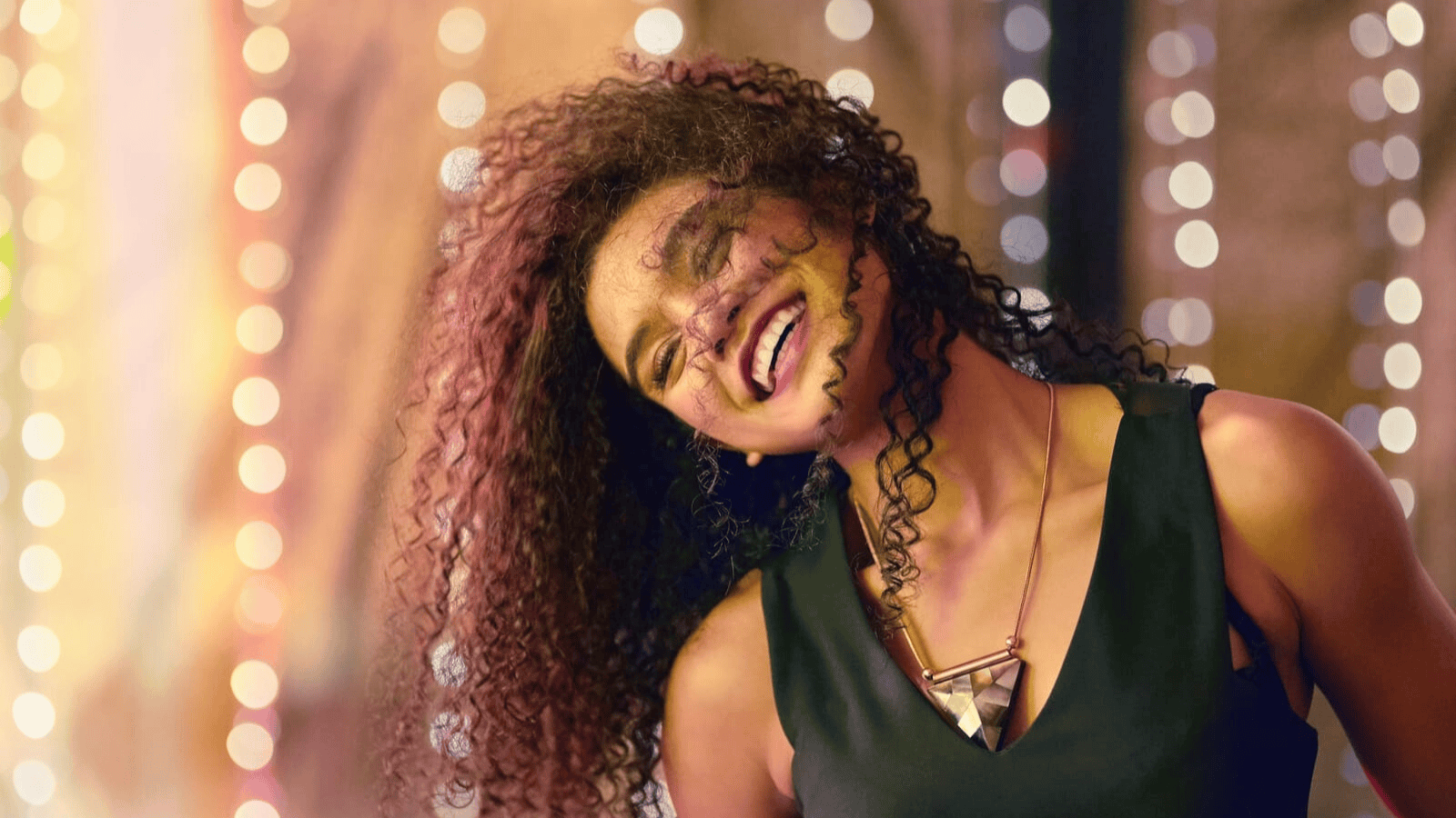 A person with curly hair is smiling with their head tilted back in front of a blurred background with bokeh lights. They are wearing a sleeveless top and a geometric necklace.