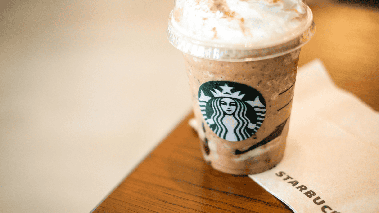 A Starbucks frappuccino topped with whipped cream sits on a wooden table next to a Starbucks-branded napkin. The cup features the Starbucks logo, and the beverage appears to be a blended iced drink with chocolate and cream.