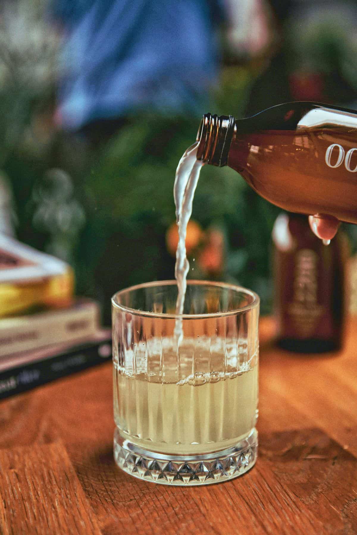 A hand pours a beverage from a brown bottle into a glass tumbler. The glass sits on a wooden surface, surrounded by a stack of books and a blurred background with green foliage.