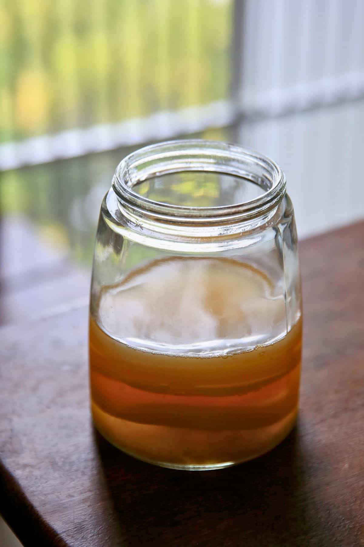 A partially filled glass jar with a gold-colored liquid, likely honey, sits on a wooden surface. A blurred window and greenery are visible in the background.