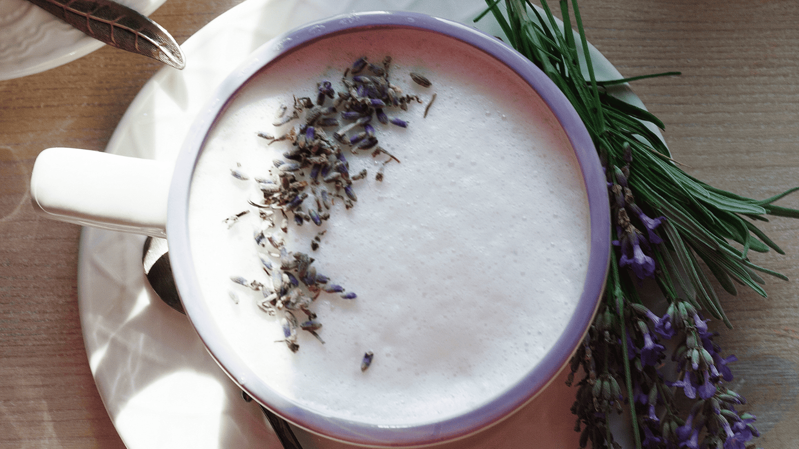 A mug of frothy milk topped with dried lavender buds sits on a white saucer with a spoon on the side. Fresh sprigs of lavender with purple flowers are placed next to the mug on a wooden surface.