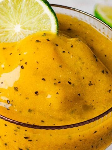 Close-up of a glass filled with a thick, yellow smoothie. The smoothie appears to have flecks of darker material blended in. A slice of lime is perched on the rim of the glass, and part of a cut passion fruit is visible in the background.