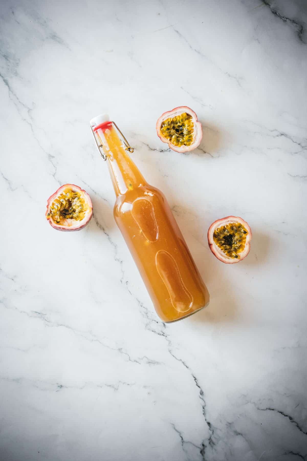 A bottle filled with a brown liquid is placed on a light marble surface. The bottle has a white cap with a red seal. There are three halves of a passion fruit arranged around the bottle, showing the yellow flesh and black seeds inside.