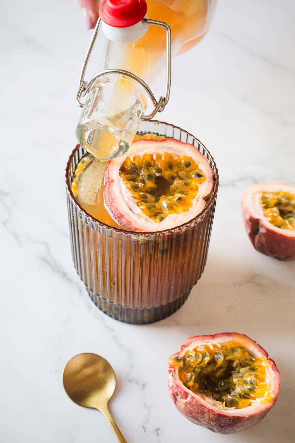 A glass with a passion fruit garnish is being filled with a drink poured from a glass bottle with a swing top. A halved passion fruit and a gold spoon are placed on a marble surface beside the glass.