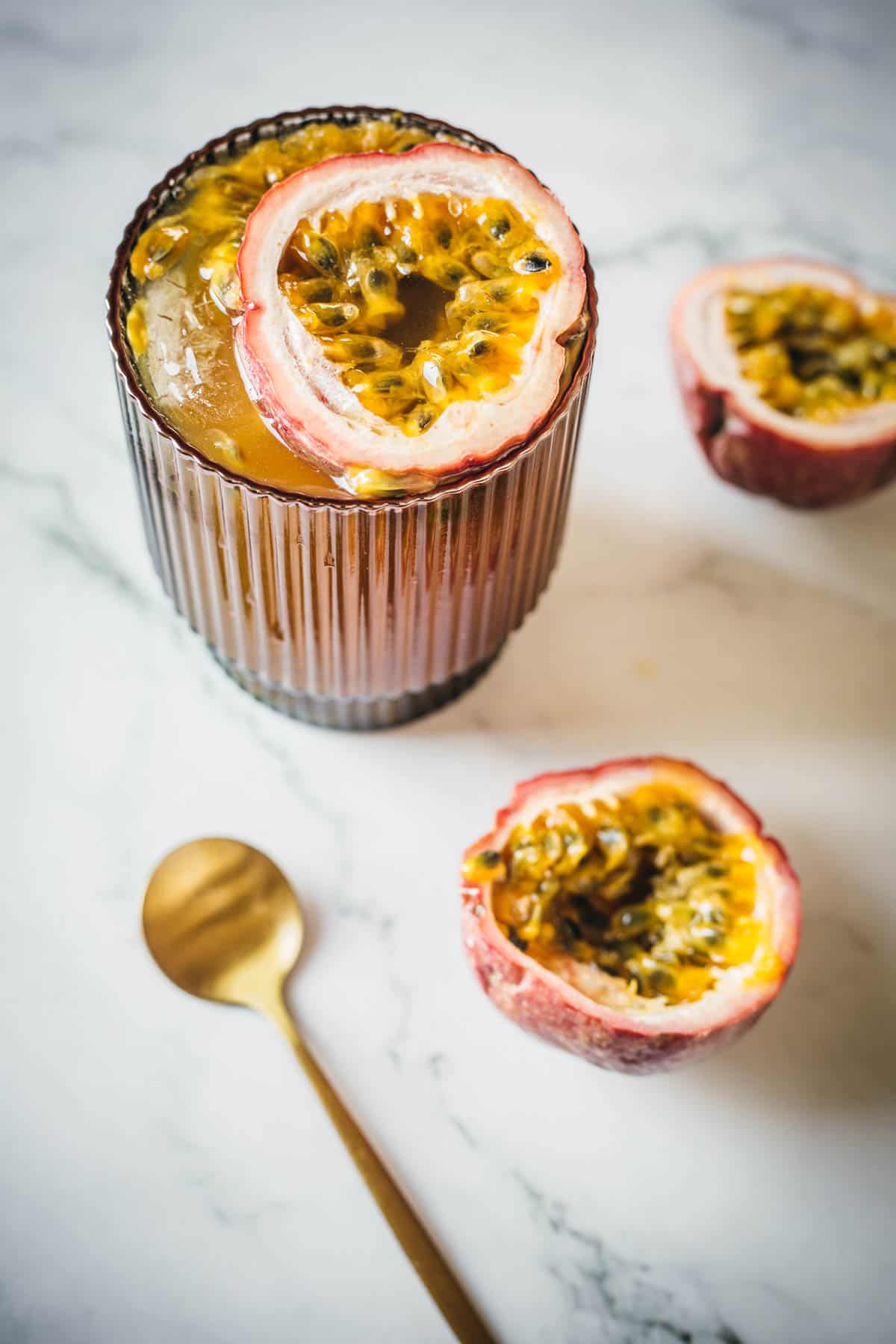 A glass of passion fruit drink sits on a marble surface, garnished with a halved passion fruit on top. Two additional halved passion fruits and a gold spoon are positioned nearby.