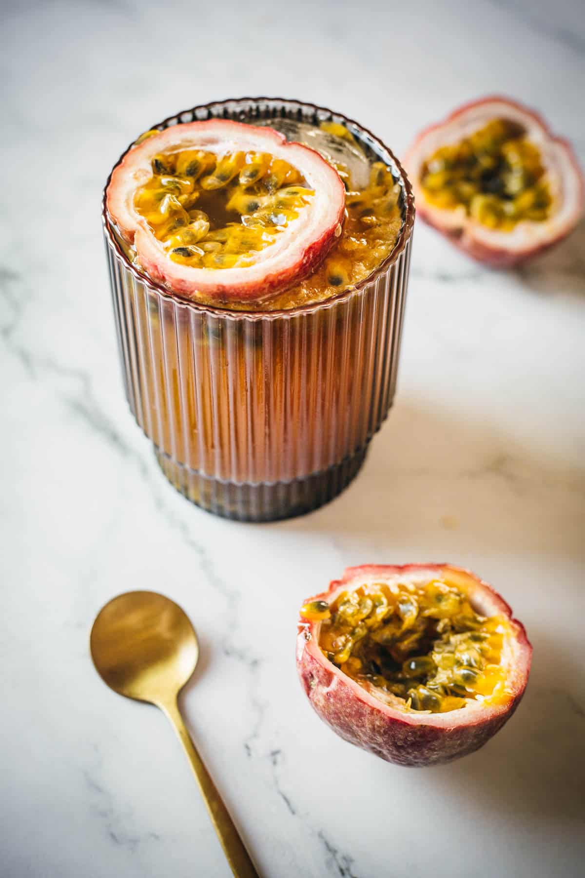 A ribbed glass filled with a passion fruit beverage sits on a marble counter. Two halved passion fruits are placed beside the glass, one on the counter and one on top of the drink. A gold spoon rests to the left of the glass.