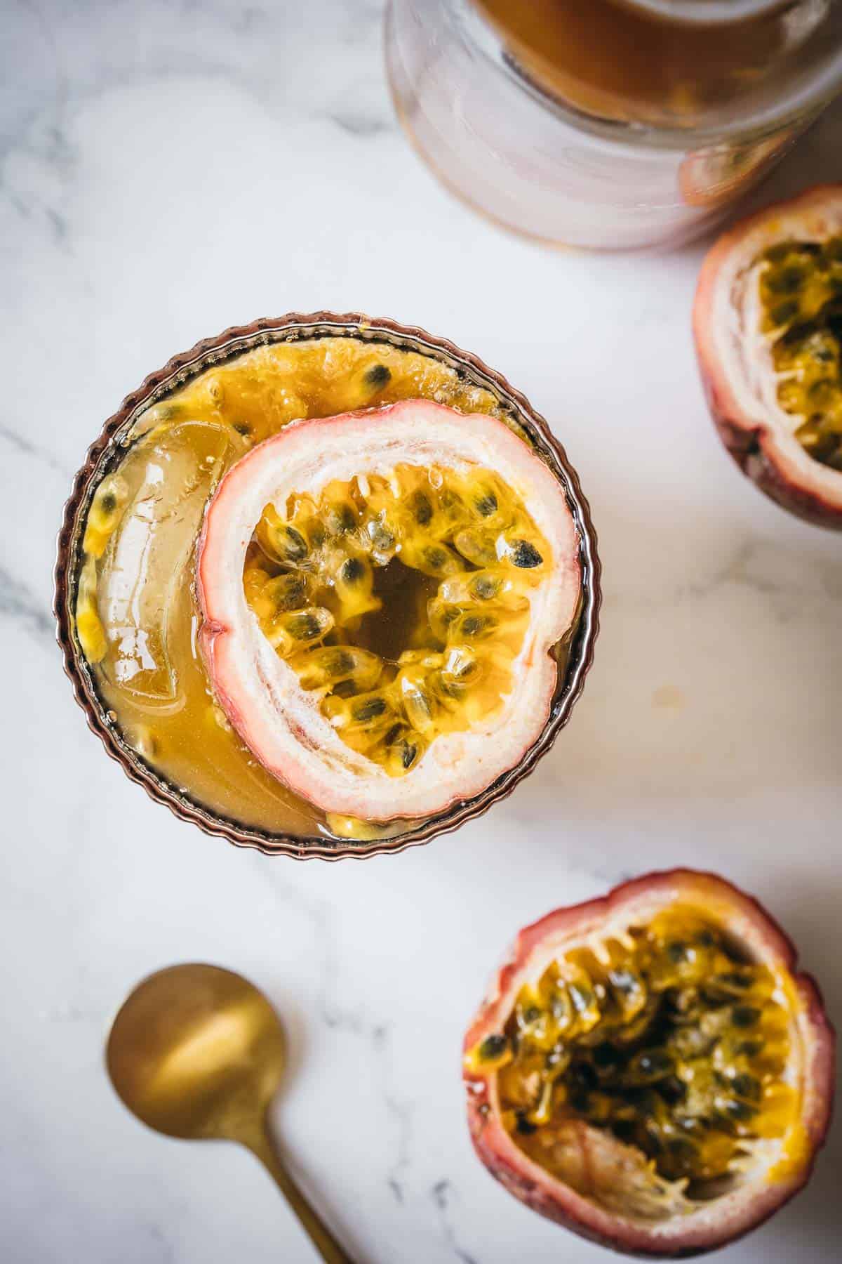 A small glass filled with a passion fruit drink, topped with halved passion fruit. Two additional halves of passion fruit and a gold spoon are placed on a marble surface beside the glass.