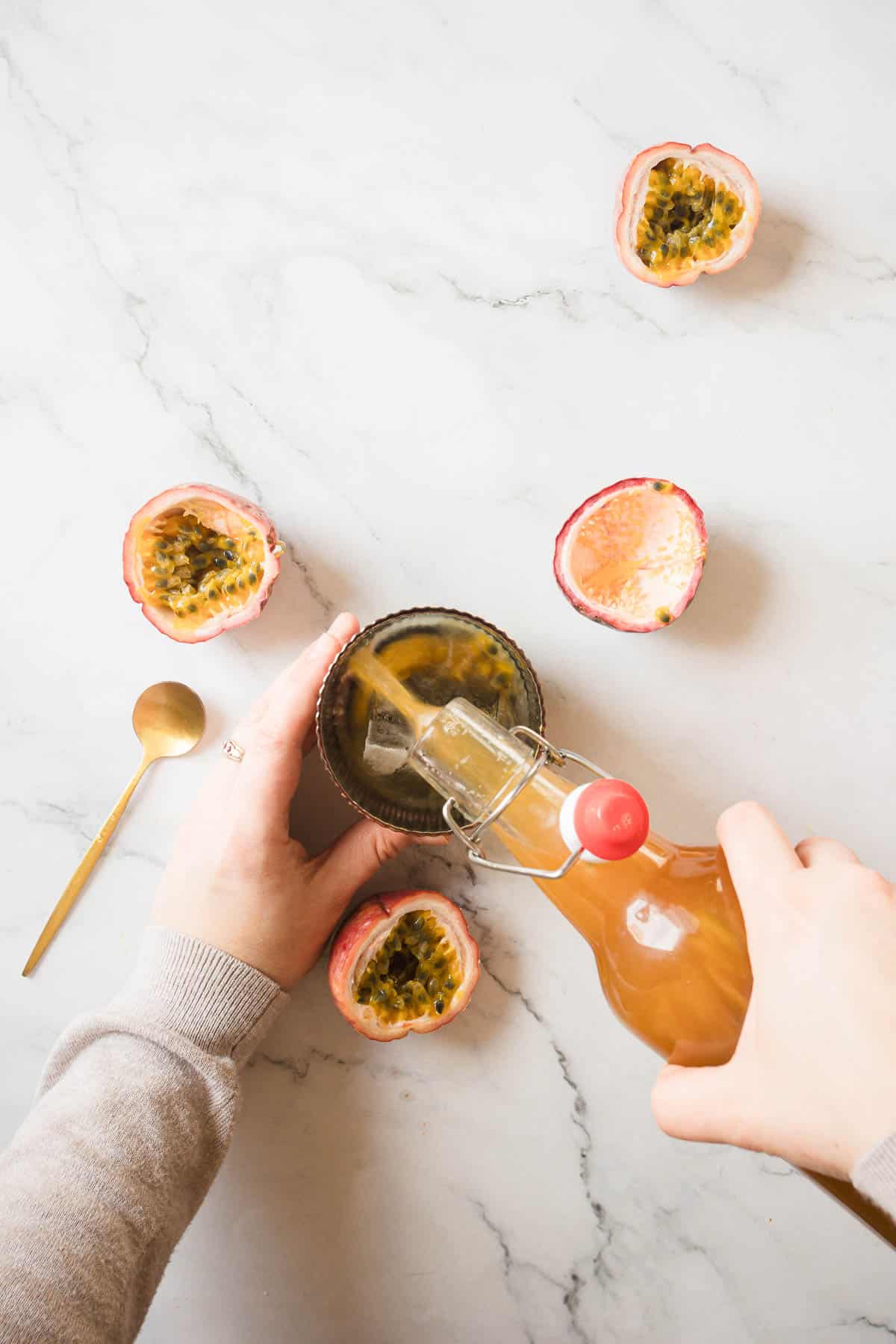 A person pours juice from a glass bottle with a red cap into a small bowl. The bowl contains passion fruit pulp. There are three half-cut passion fruits and a gold spoon placed on a white marble surface around the bowl.