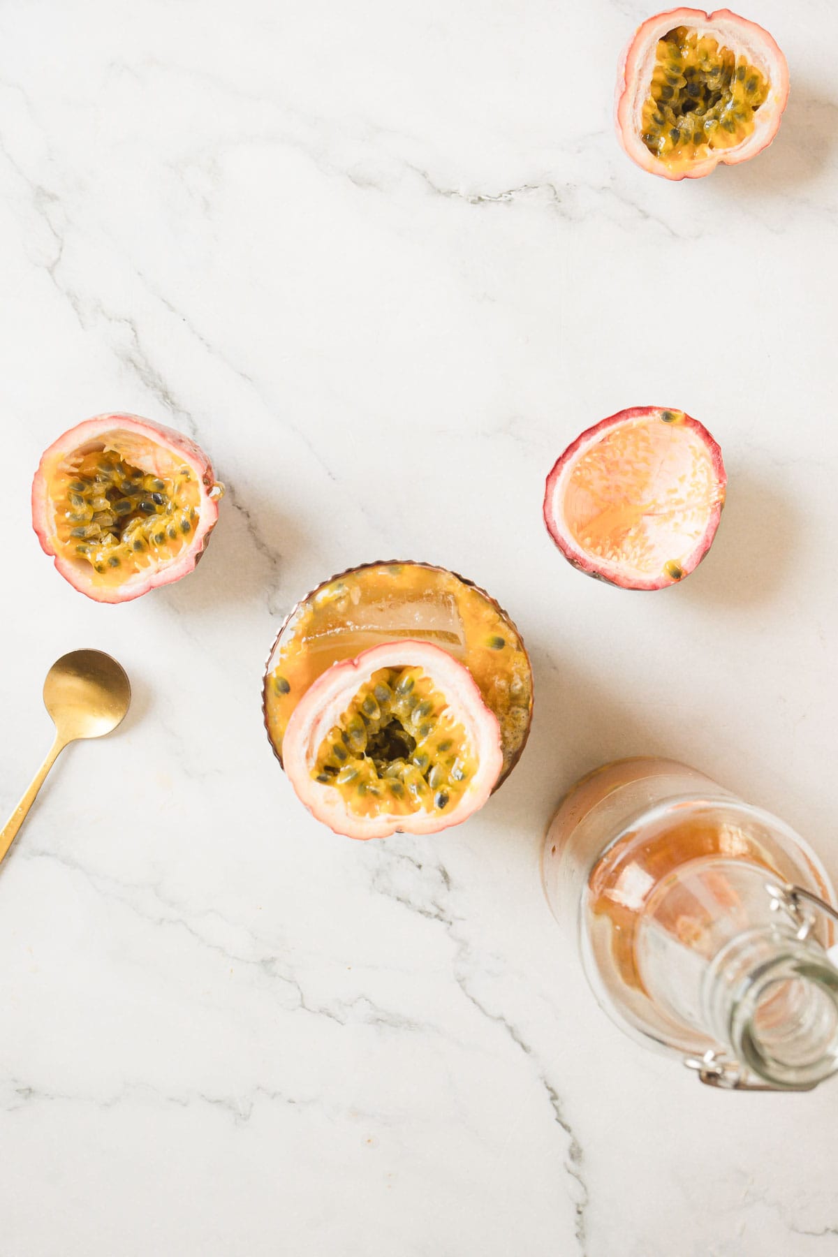 A marble surface with a halved passion fruit on the left side, two halved passion fruits in the middle of a glass cup filled with passion fruit juice, a gold spoon next to the cup, and a glass bottle with juice on the right side.