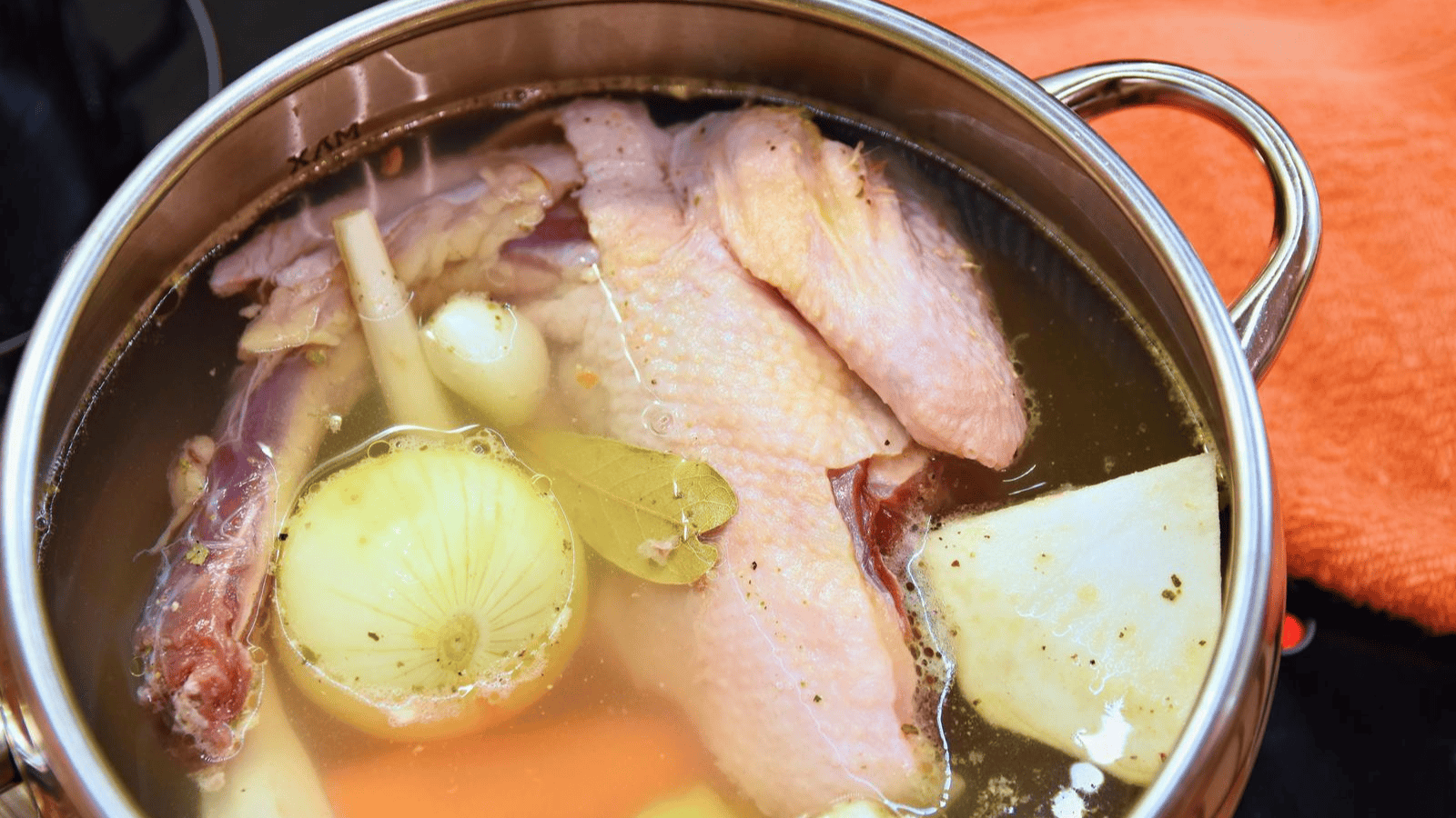 A large pot filled with poultry parts and vegetables, including onions, carrots, and a bay leaf, submerged in water. The pot is stainless steel with an orange cloth in the background. The contents appear to be cooking or simmering.