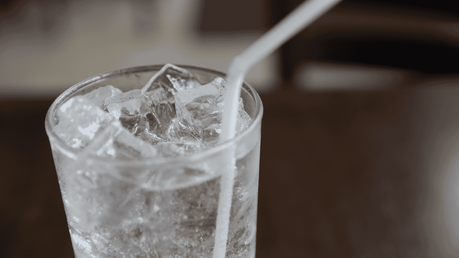 A close-up image of a clear glass filled with ice cubes and water. A white plastic straw is leaning against the inside edge of the glass. The background is blurred and appears to have a brown table and chairs.
