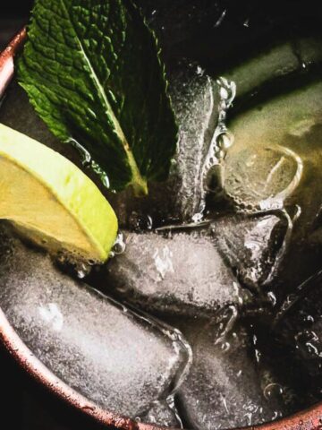 Close-up image of a moscow mule cocktail in a copper mug, garnished with a lime wedge and fresh mint leaves, filled with ice cubes.