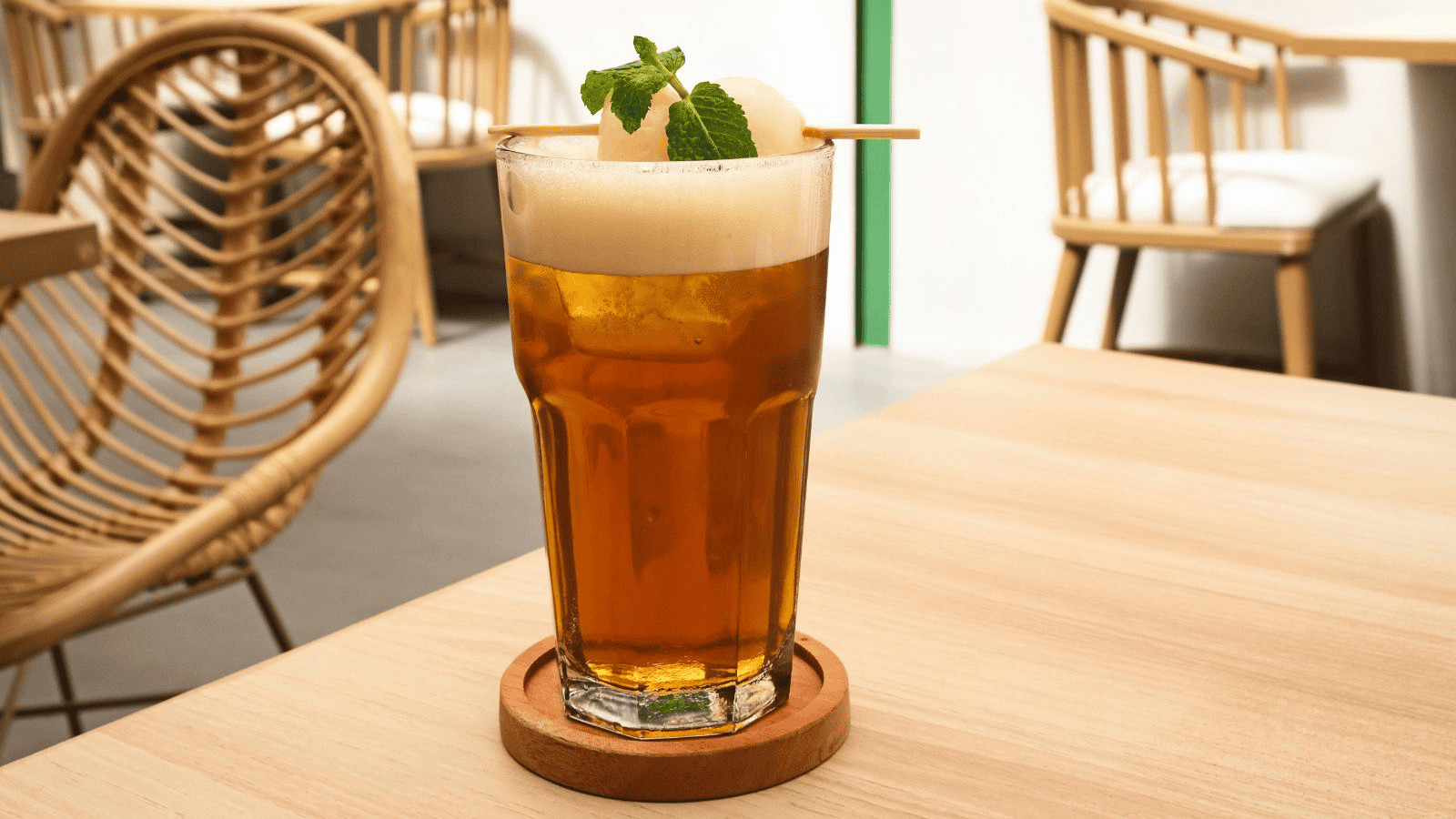 A tall glass of iced tea with foam sits on a wooden table in a cafe. The drink is garnished with a scoop of sorbet and a sprig of mint, served on a round wooden coaster with light-colored wooden chairs in the background.