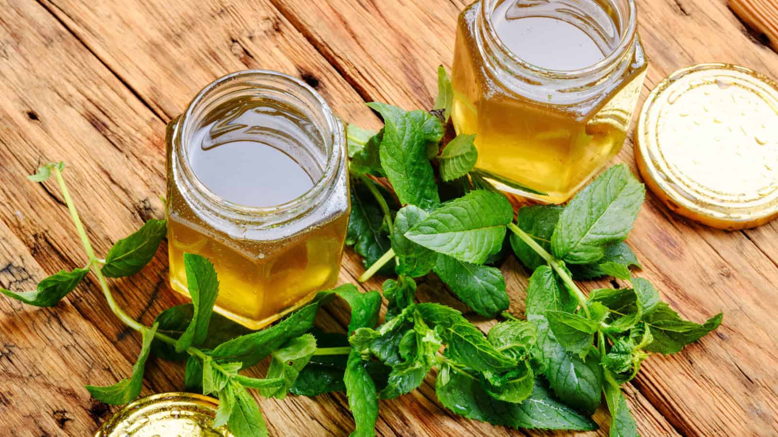 Two hexagonal glass jars filled with golden honey are placed on a wooden surface. Each jar has a gold lid lying beside it. Fresh green mint leaves are scattered around the jars.