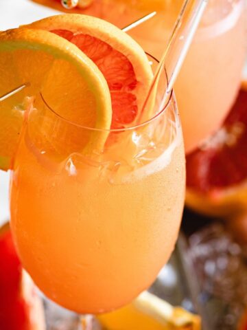 A refreshing citrus cocktail in a stemmed glass, garnished with orange and grapefruit slices. the drink is served on a tray with whole and halved citrus fruits.