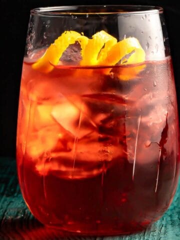 A glass of iced tea with a lemon twist garnish sits on a turquoise wooden table, illuminated by subtle backlighting, highlighting its vibrant red color and condensation on the glass.