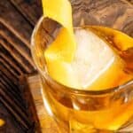 A glass of whiskey with ice and a lemon peel garnish, sitting on a wooden coaster beside a cut orange on a rustic wooden table.