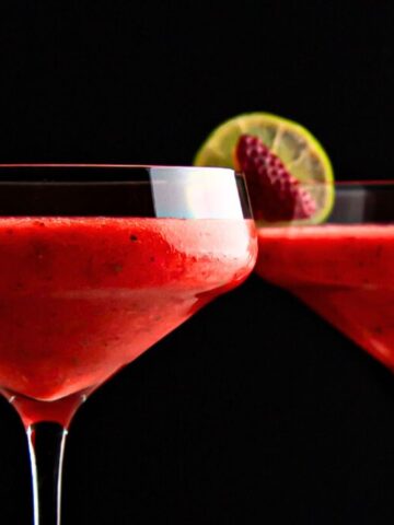 Two glasses of strawberry daiquiri garnished with lime slices and strawberries, set against a dark background.