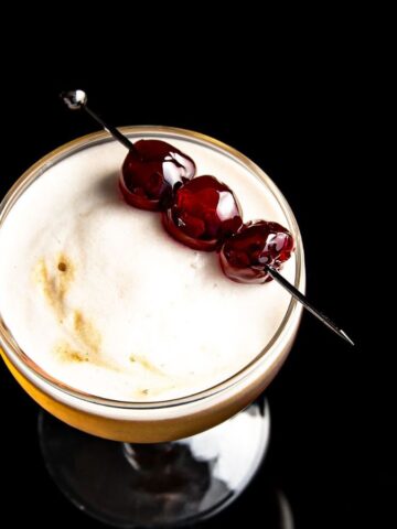 A close-up of a cocktail glass against a black background, containing a creamy, frothy beverage garnished with three cherries on a skewer.