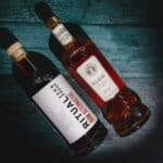 Two bottles of non-alcoholic spirits lie on a dark green wooden surface. One bottle, labeled "Ritual Zero Proof," is clear with a white label and a wooden cap. The other bottle, labeled "Fluère," is amber and has an intricately designed label and cap.