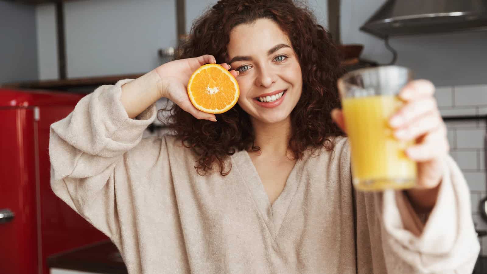A person with curly hair and wearing a bathrobe smiles in a kitchen while holding a glass of orange juice in one hand and a sliced orange in the other. A red refrigerator and white-tiled backsplash are visible in the background.