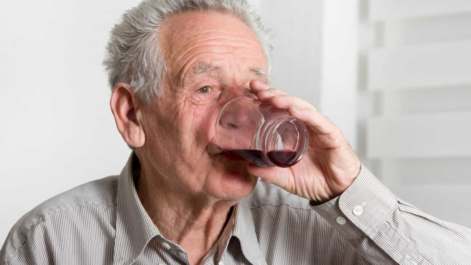 An elderly man with gray hair is drinking wine from a clear glass. He is wearing a striped button-up shirt and is indoors with light-colored walls and blinds in the background.