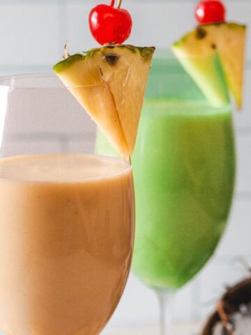 Two colorful smoothies in clear glasses, one orange and one green, each garnished with a pineapple slice and cherry, displayed against a tiled backsplash.