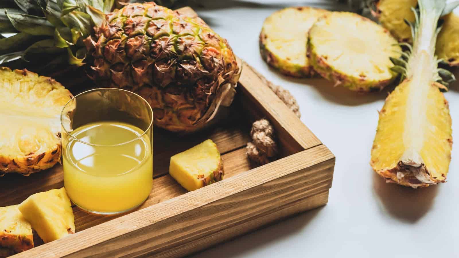A wooden tray containing a whole pineapple, sliced pineapple, and a glass of pineapple juice. Additional pineapple slices are on the table beside the tray. Sunlight illuminates the scene, highlighting the freshness of the fruit.