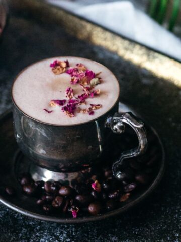 A frothy beverage in a vintage metal cup, adorned with pink petals on top, presented on a dark tray with coffee beans and cardamom pods. a rustic setup with soft lighting and a moody ambiance.