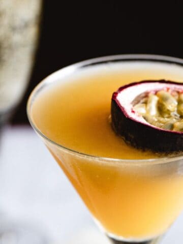 A close-up of a passion fruit cocktail in a martini glass, garnished with a half passion fruit. a blurred champagne glass is in the background on a dark counter.