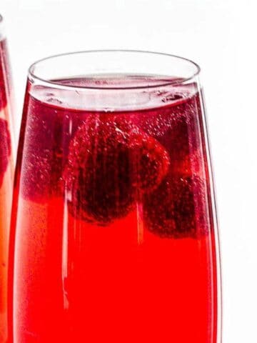 Two glasses of red sparkling beverage with raspberries floating in them, set against a bright white background.
