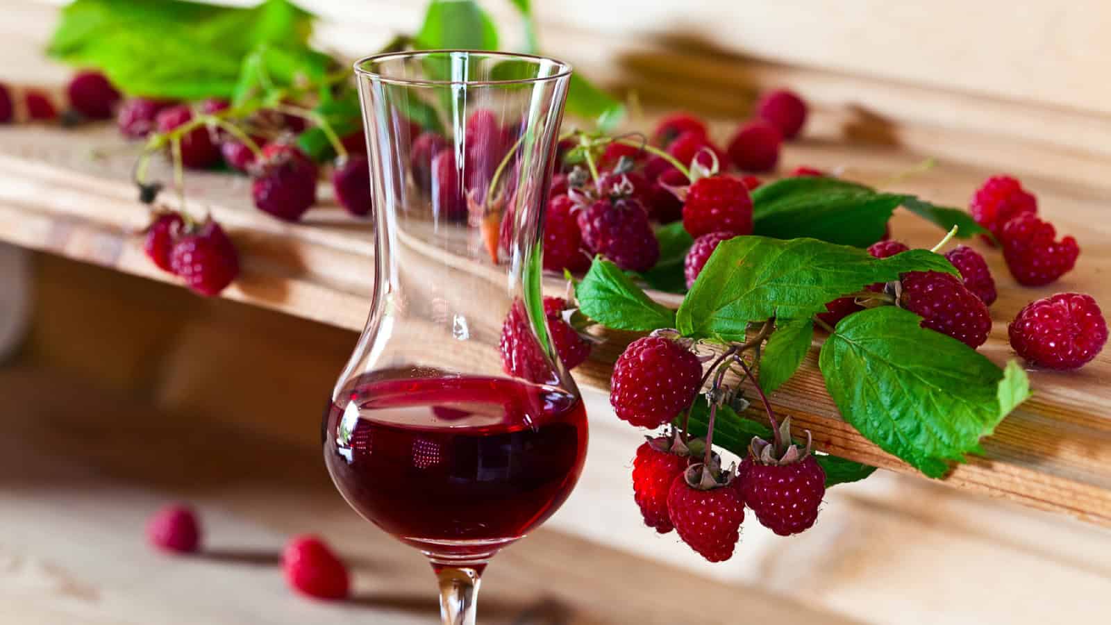 A glass of red wine is placed on a wooden surface next to a bunch of fresh raspberries and green leaves. Raspberries are scattered around, and a few are still attached to the stems. The background is a rustic wooden setting.
