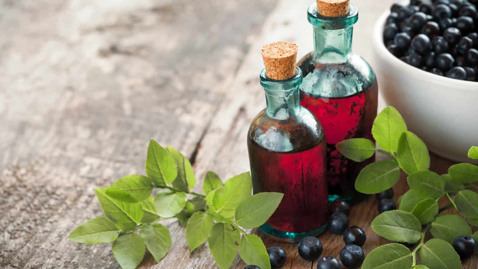 Two small glass bottles with cork stoppers containing a red liquid are placed on a wooden surface. Next to the bottles are green leaves and scattered blueberries, with additional blueberries in a white bowl.