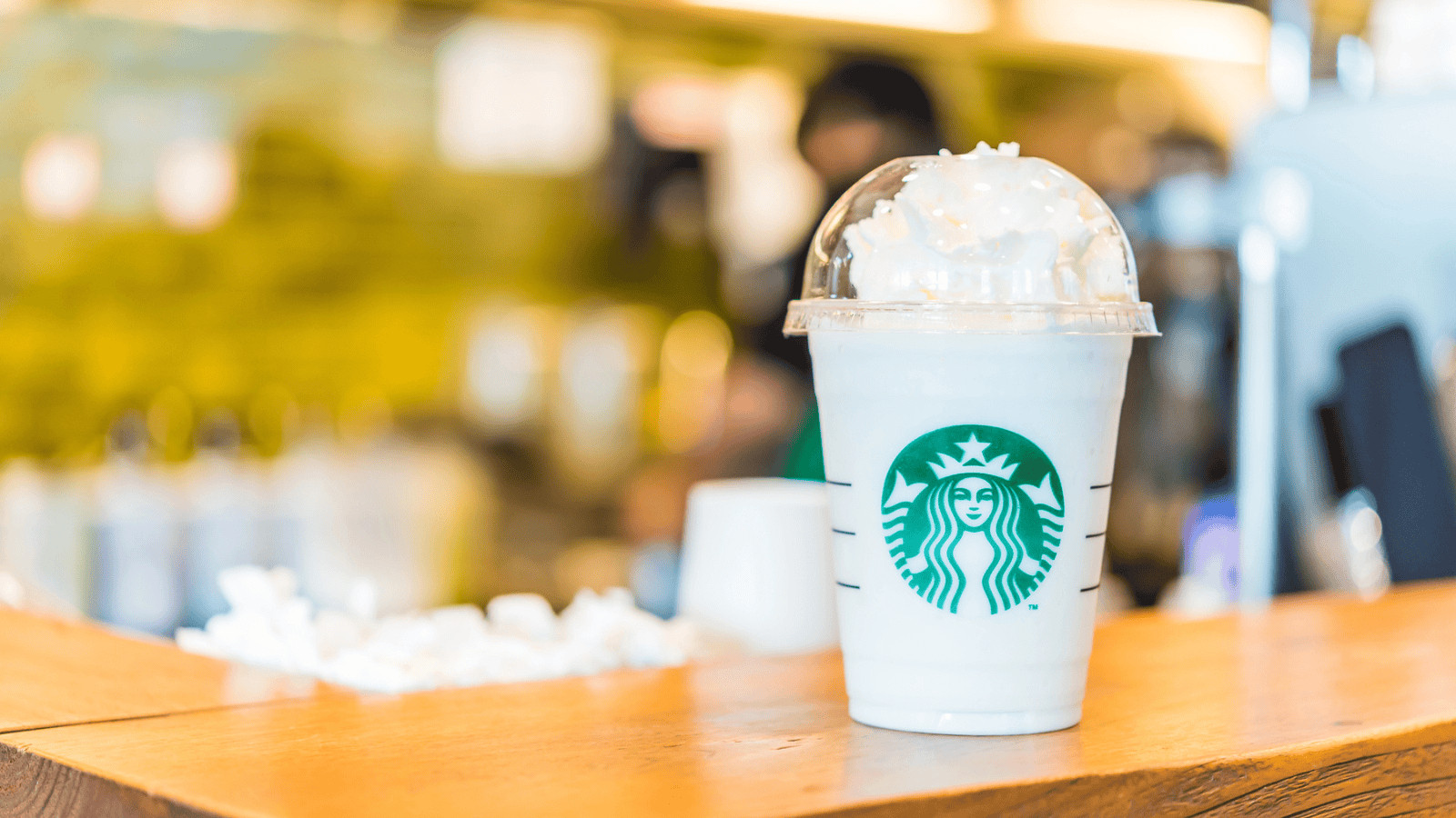 A Starbucks Frappuccino topped with whipped cream in a clear plastic cup sits on a wooden counter in a coffee shop. The iconic green Starbucks logo is visible on the cup. The background consists of blurred tables, cups, and a barista.