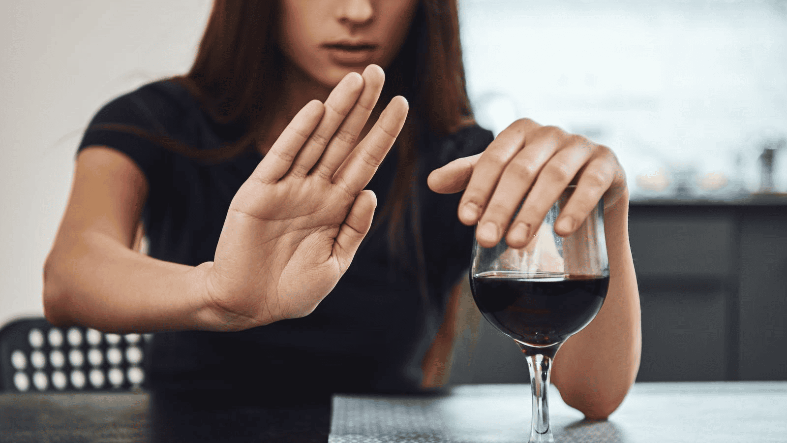 A person in a dark shirt extends their right hand to signal "stop" while their left hand slightly covers a glass of red wine on a table. The background shows a blurred kitchen.