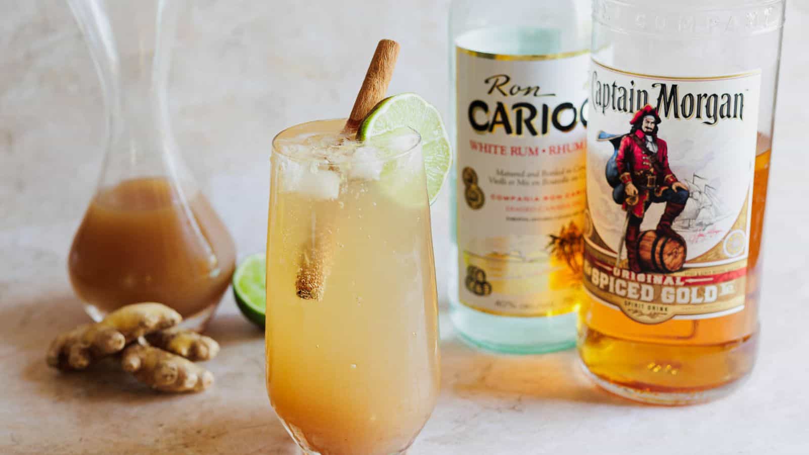 A glass of iced cocktail with a lime wedge and cinnamon stick, alongside bottles of ron cario white rum and captain morgan spiced gold rum, with ginger roots nearby.