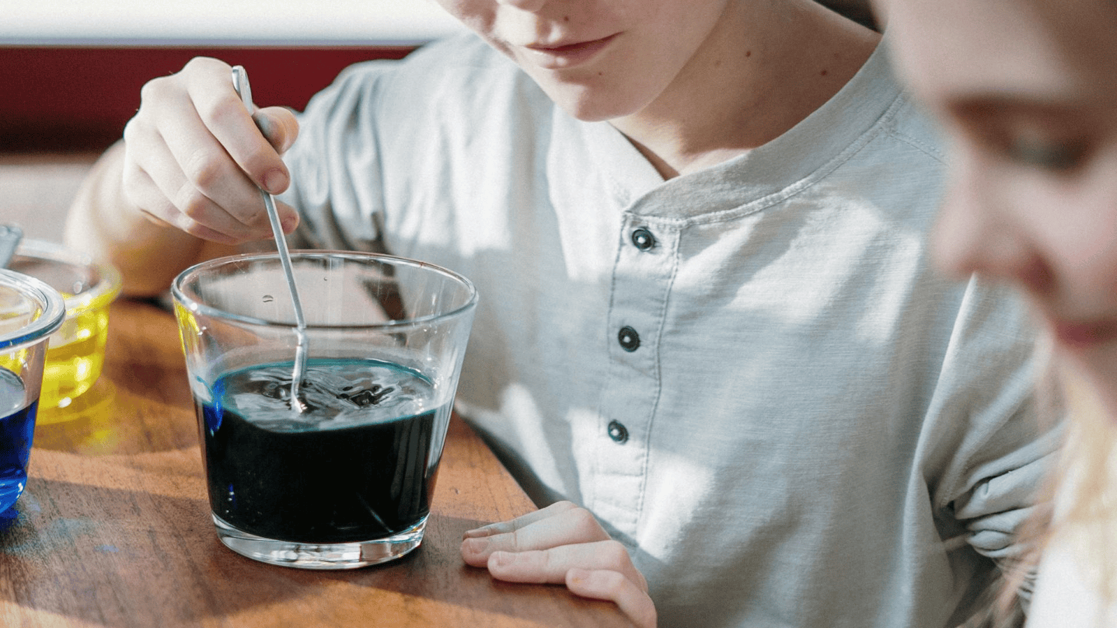 A person in a light grey shirt is stirring liquid with a spoon in a glass containing dark blue liquid, on a wooden table. Additionally, three other containers with yellow and blue liquid are partially visible on the table.