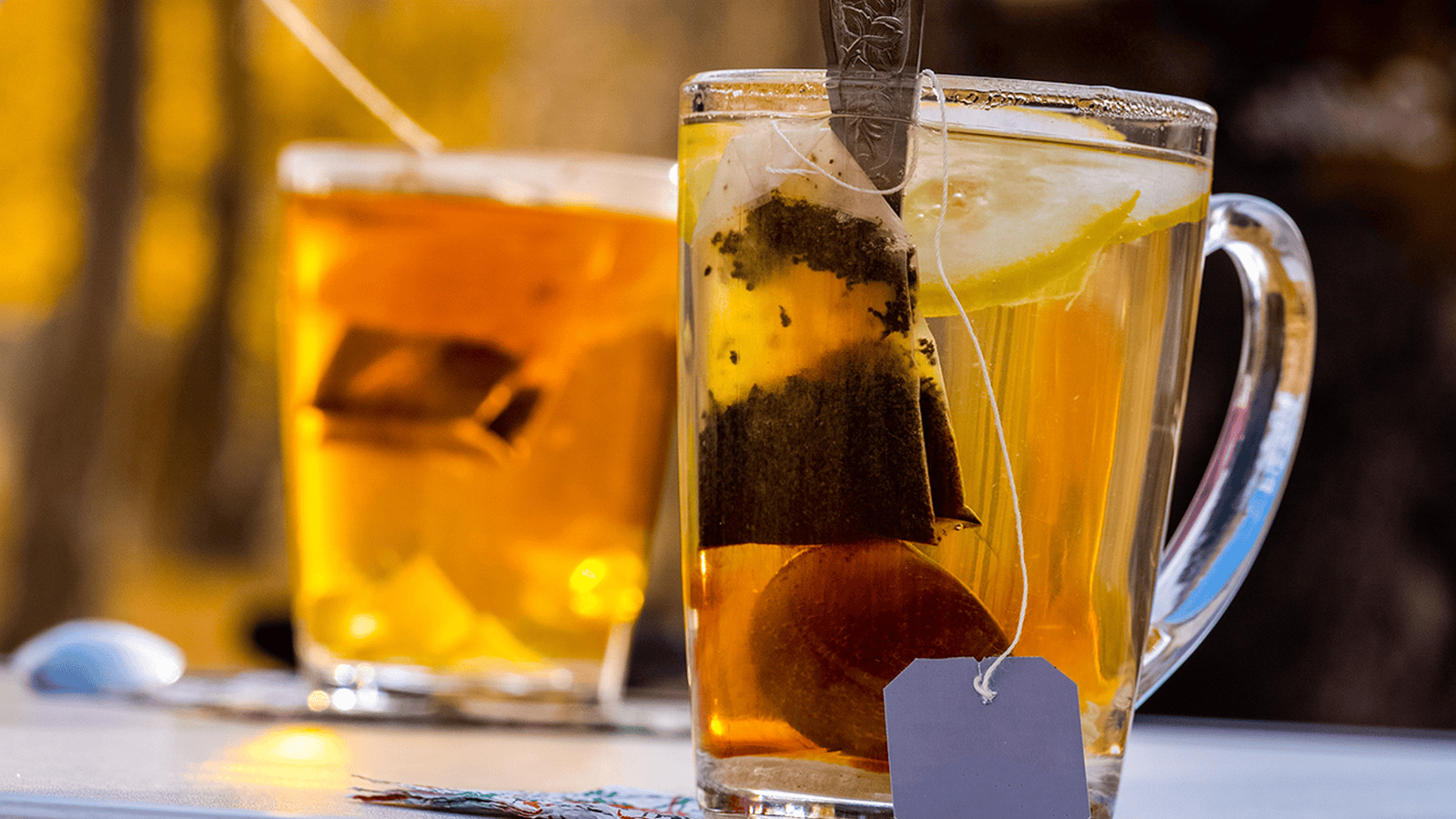 Two clear glass mugs of tea with tea bags and lemon slices inside are placed on a table. One mug is in the foreground with a tea tag visible, and the other is in the background. The sunlight creates a warm glow on the tea and the surrounding area.
