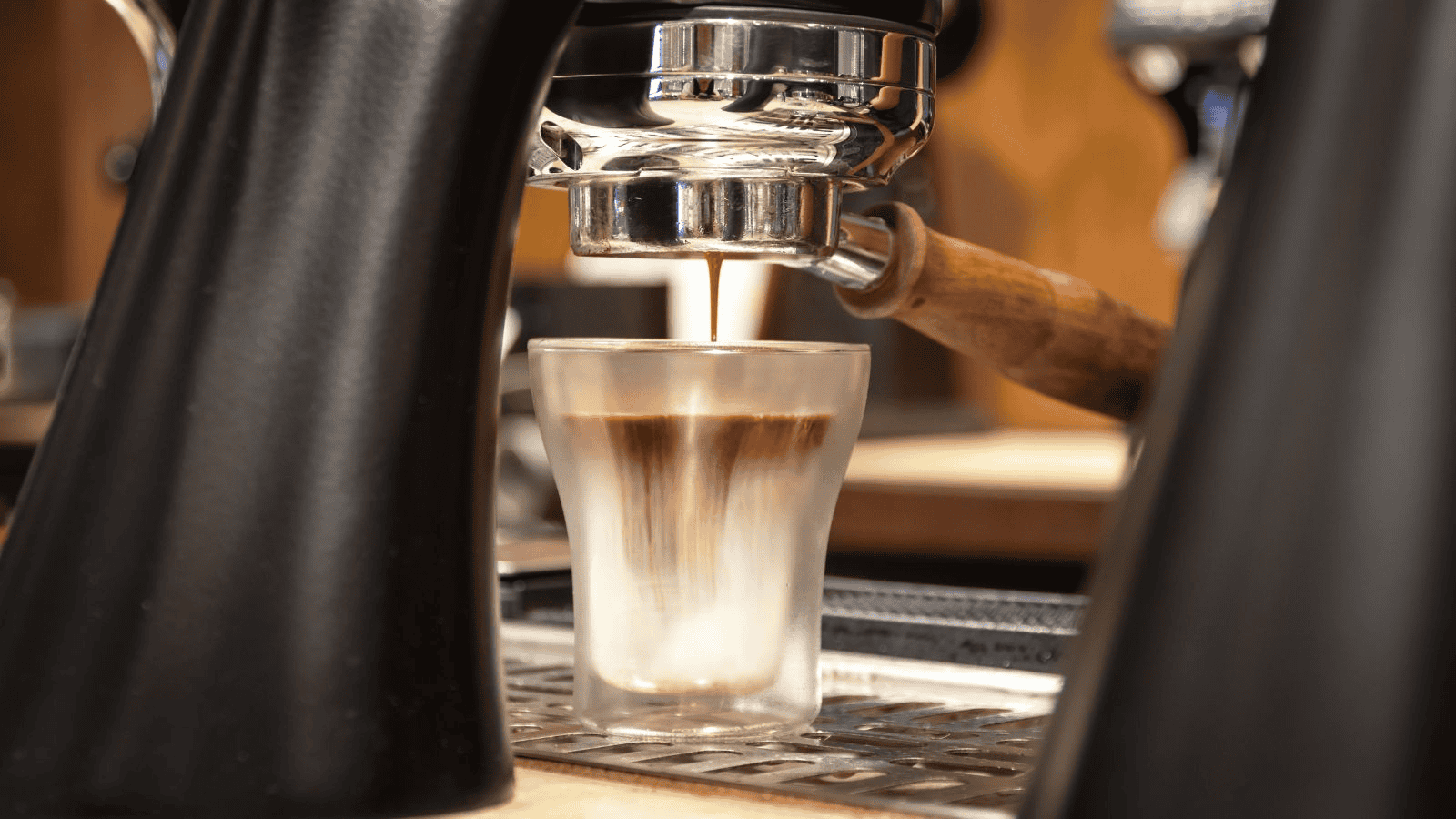 A close-up image of a coffee machine dispensing espresso into a glass espresso shot cup. The espresso appears to be mixing with milk, creating a layered effect. The machine has a wooden handle, and the background is slightly blurred..