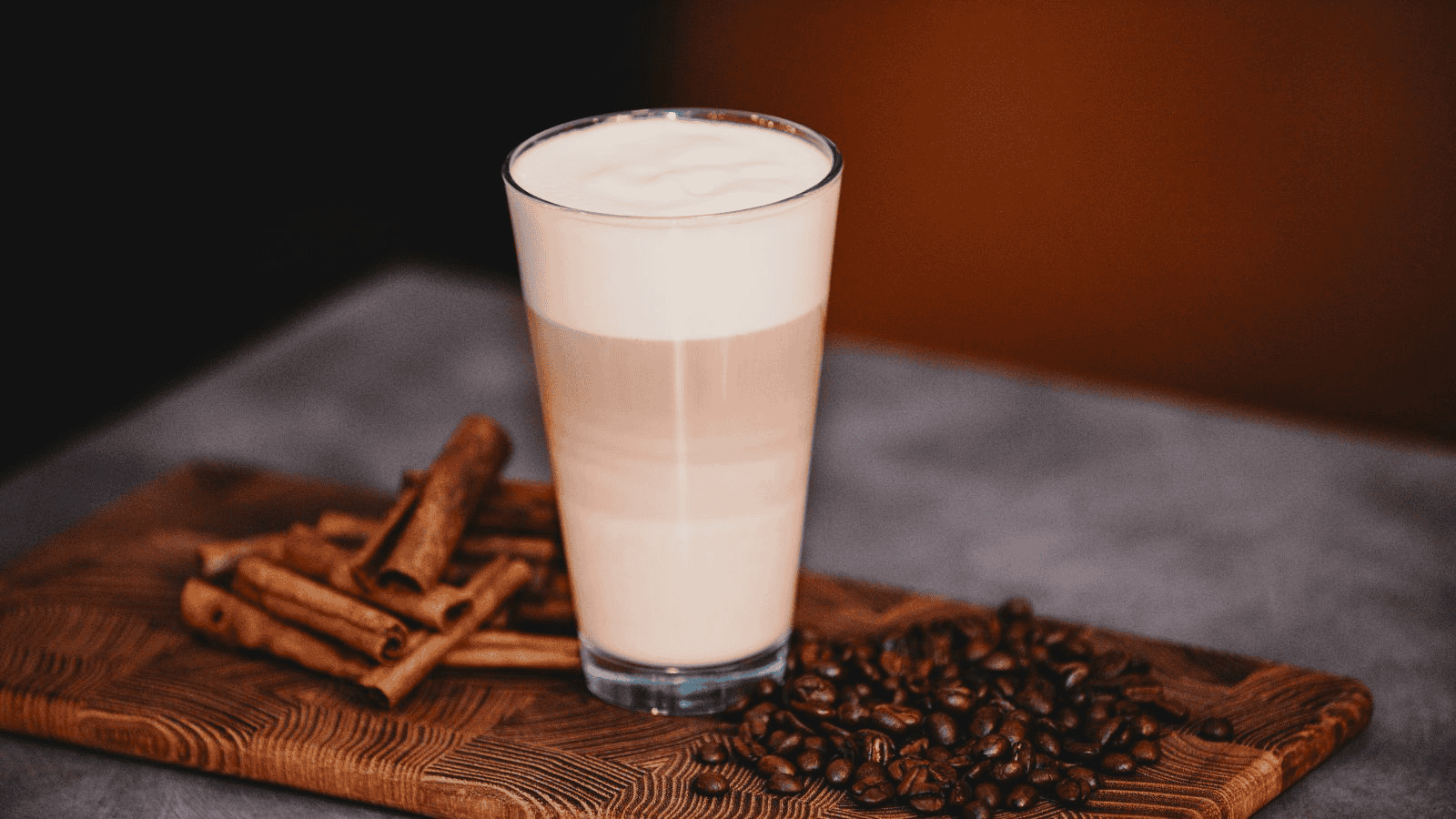A layered latte topped with foam sits on a wooden cutting board. Surrounding the glass are a few cinnamon sticks and scattered coffee beans. The background is blurred, emphasizing the drink and ingredients.
