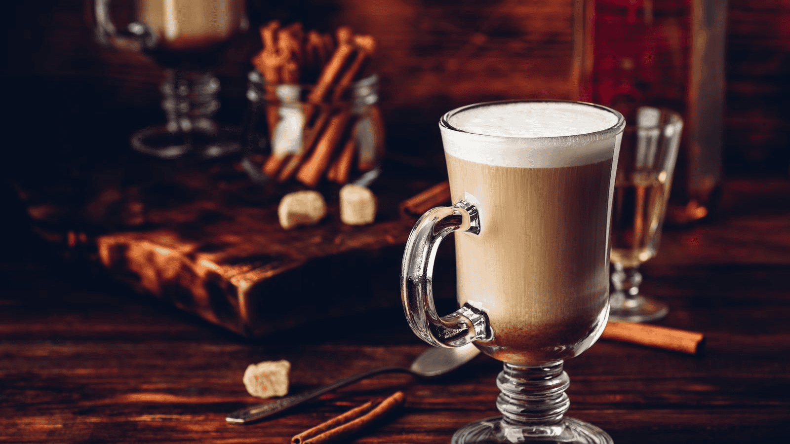 A glass of creamy hot beverage, possibly coffee or latte, on a wooden surface with a jar of cinnamon sticks, sugar cubes, and a metal fork in the background. Another glass with a yellowish liquid is partially visible on the right side.