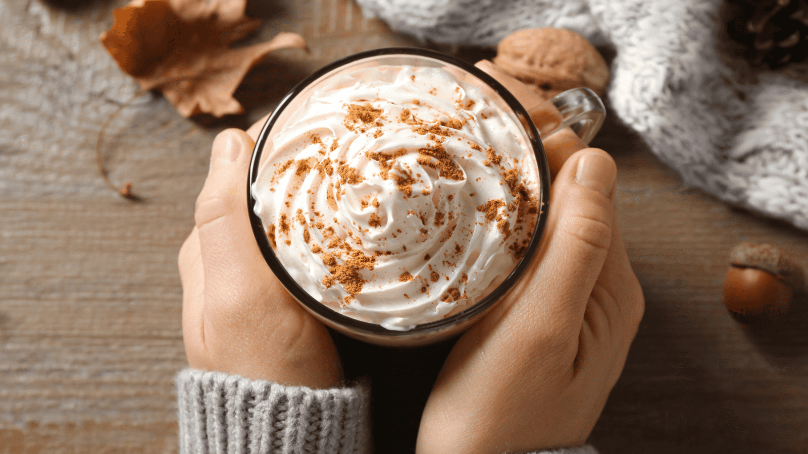 A person holding a clear mug filled with a hot beverage topped with whipped cream and sprinkled with cinnamon. The scene includes a knitted fabric, leaf, walnut, and acorn on a wooden surface.