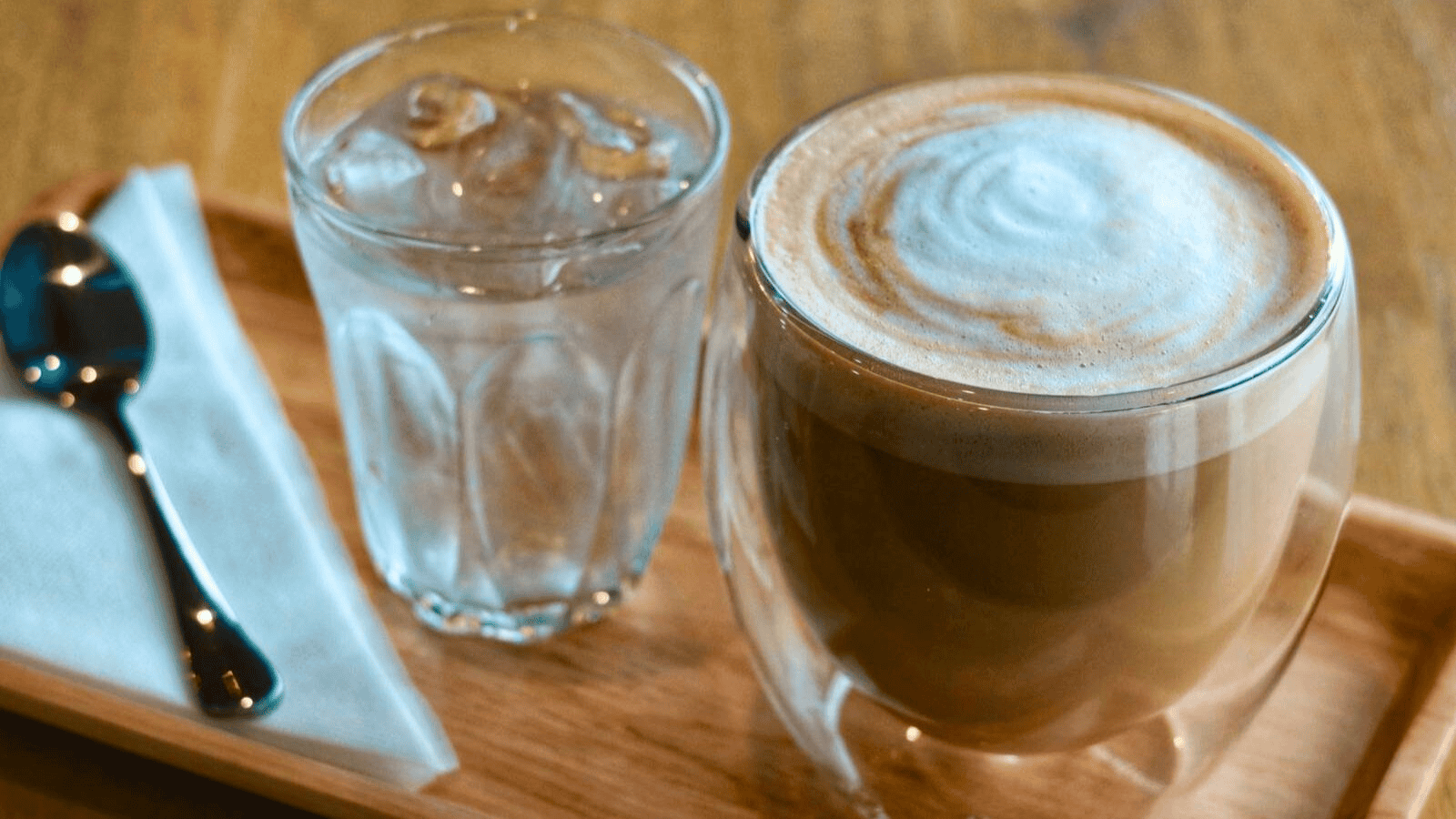 A wooden tray holds a glass of iced water, a cup of foamy coffee, a metal spoon, and a white napkin. The coffee has a light layer of foam on top and is in a double-walled glass. The tray is set on a wooden surface.