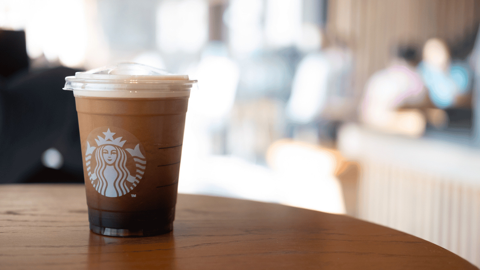 A plastic cup of iced coffee sits on a wooden table in a caf&eacute;. The cup has the Starbucks logo on it and contains a dark coffee base with a creamy top layer. The background is blurred, with out-of-focus people and indoor seating.