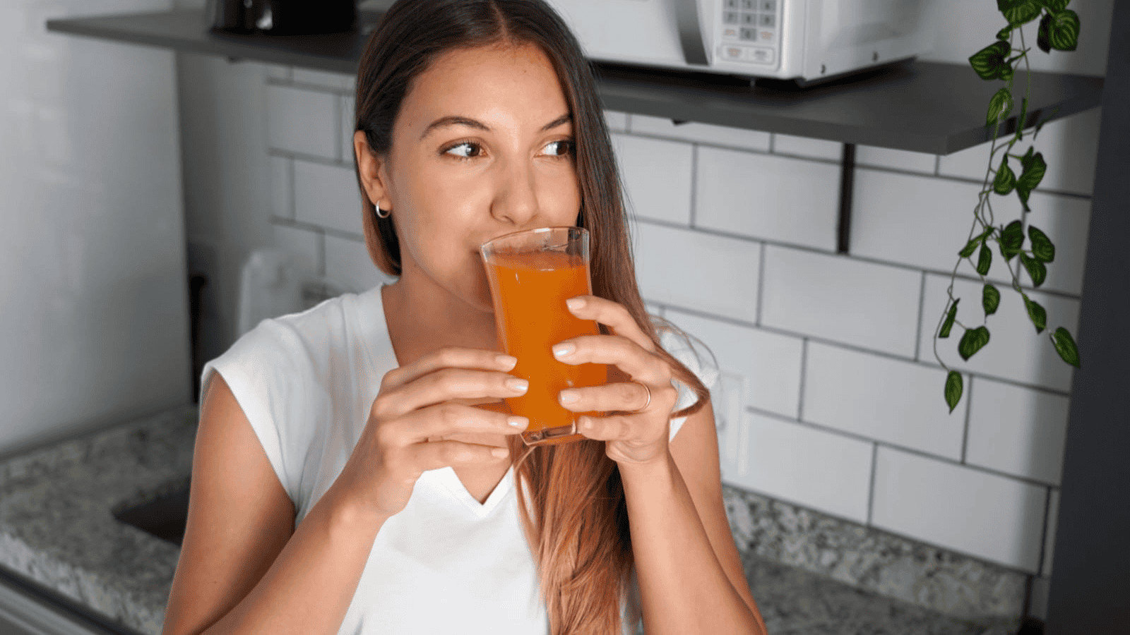 A woman with long hair is drinking orange juice from a glass in a modern kitchen. She is wearing a white shirt and looking to the side. The background includes a shelf with a microwave and some green plants.